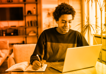 Young boy smiling while facing a laptop and writing with a pen in a journal next to the laptop. Warm red-yellow gradient overlay.
