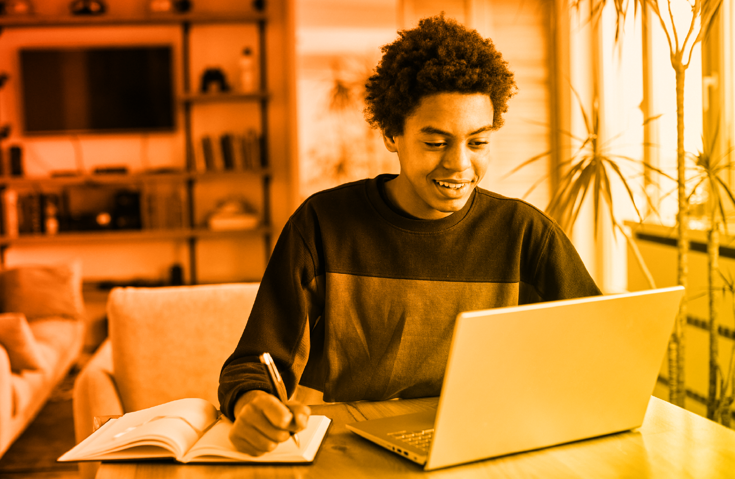 Young boy smiling while facing a laptop and writing with a pen in a journal next to the laptop. Warm red-yellow gradient overlay.